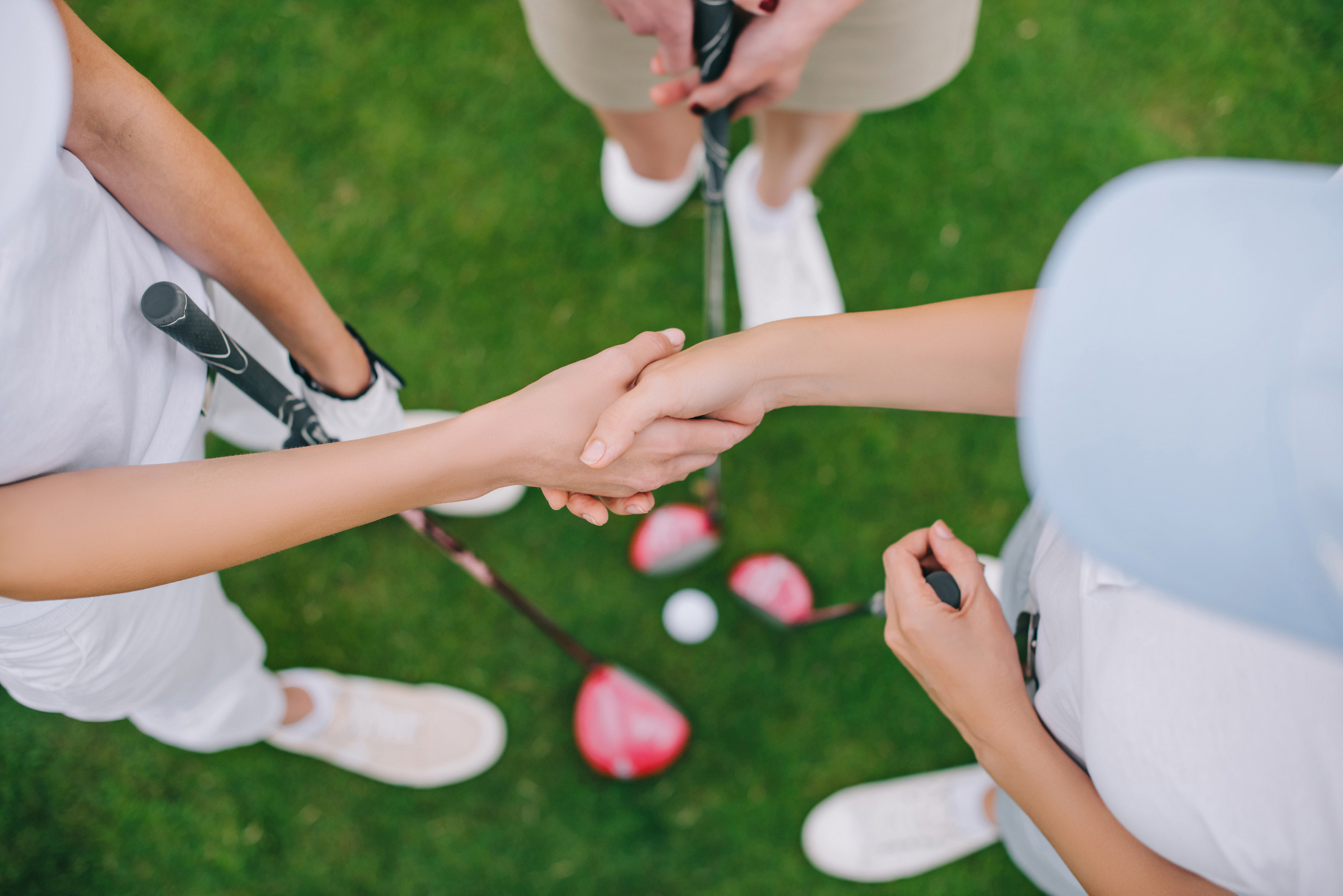 overhead view of female golf players with golf clubs shaking hands while standing on green lawn